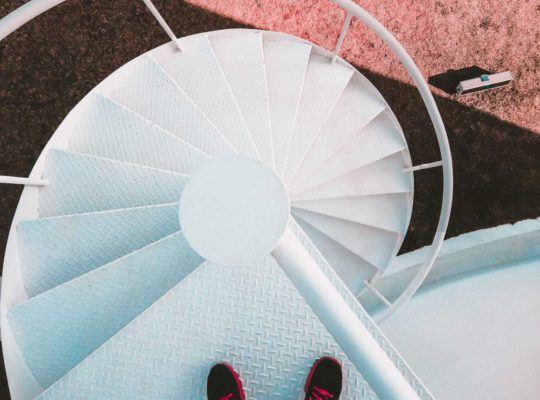 Blick nach unten auf ein paar Füße, die in schwarz-roten Schuhen auf einer Wendeltreppe stehen